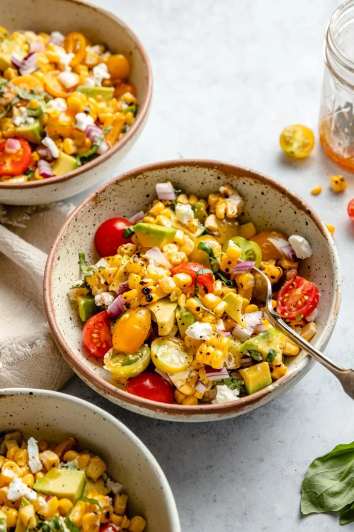 three bowls showing a grilled corn salad with sliced cherry tomatoes, goat cheese, and avocado with a hot honey vinaigrette from Ambitious Kitchen