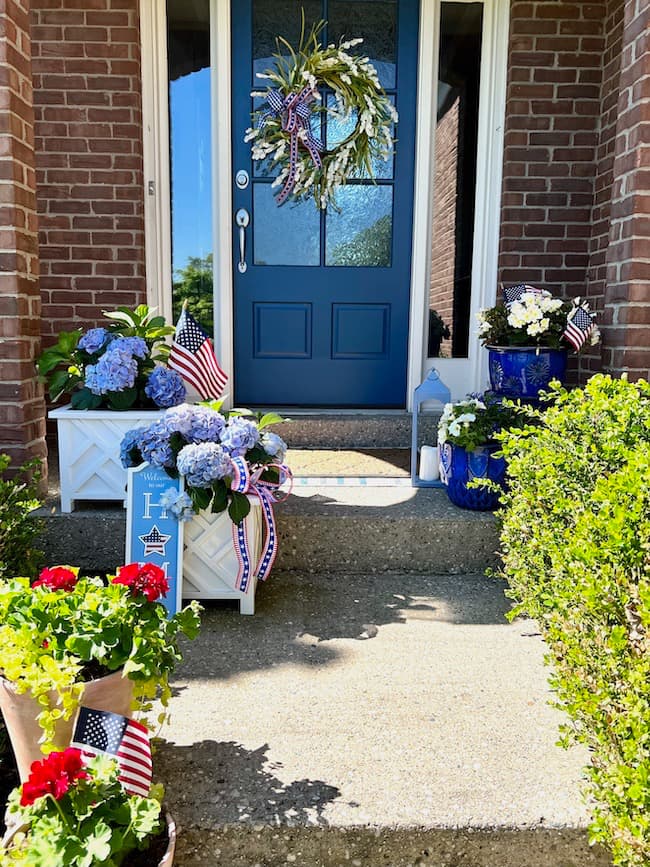 patriotic front porch with a blue front door. on the door is white wreath with a patriotic ribbon. There are blue hydrangeas in white box planters on the front porch steps. There are red geraniums planted in other planters.