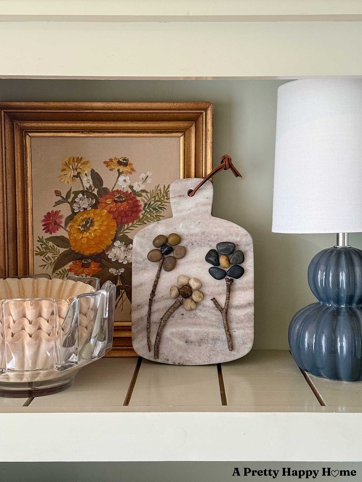 DIY pebble flower art on a marble cutting board canvas shown in front of a painting of flowers and next to a small blue lamp and a crystal bowl of coffee filters