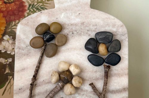 pebble flower art made by glueing small pebbles in the shape of flower petals and attaching a stem made from a stick and these are all glued onto a small marble cutting board