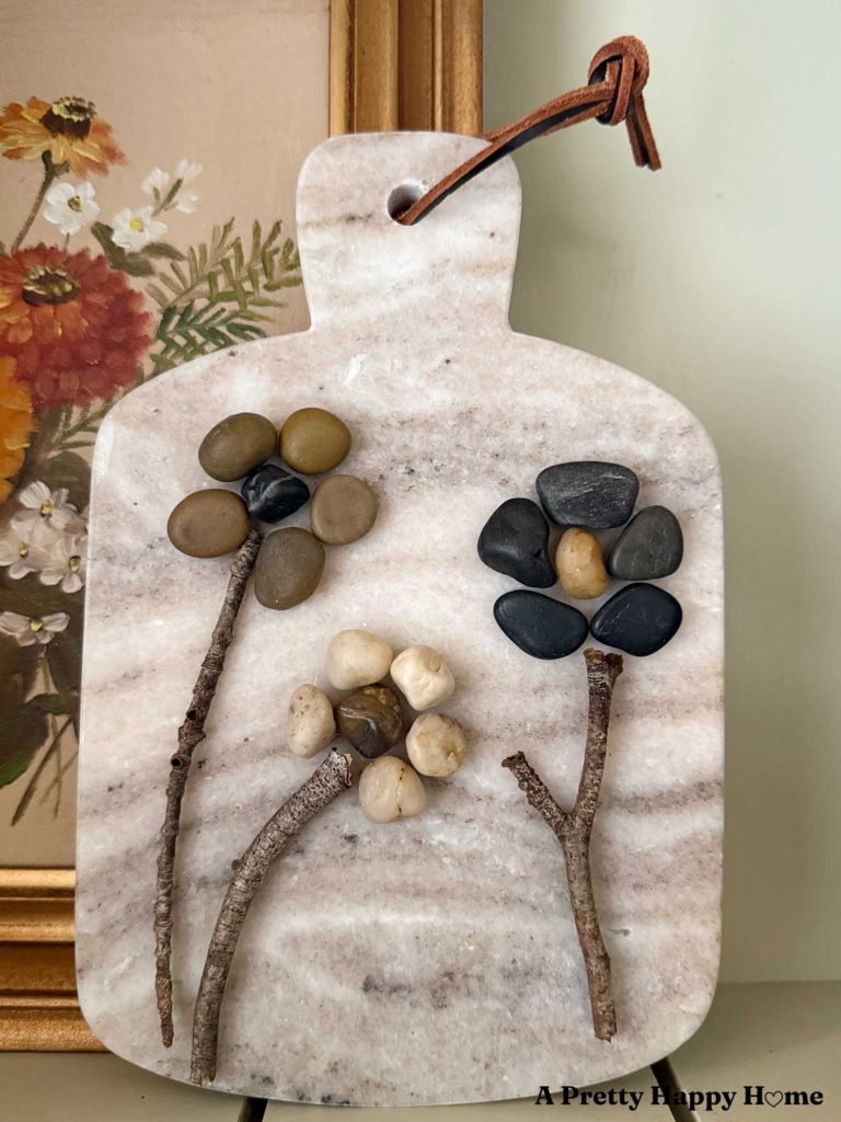 pebble flower art made by glueing small pebbles in the shape of flower petals and attaching a stem made from a stick and these are all glued onto a small marble cutting board
