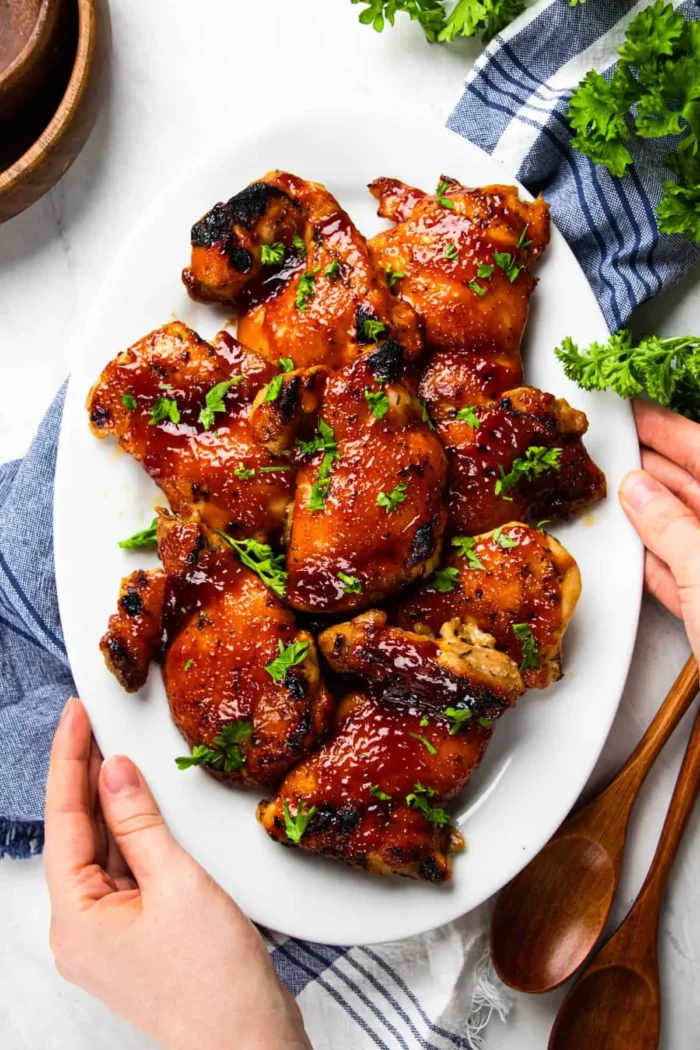 woman's hands holding a white plate full of baked bbq boneless skinless chicken thighs garnished with a sprinkle of parsley