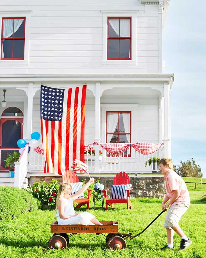 a child pulling another in a red radio flyer wagon. Behind them is an old white house with red windows that's decorated for 4th of July with an american flag and red gingham sashes on the porch