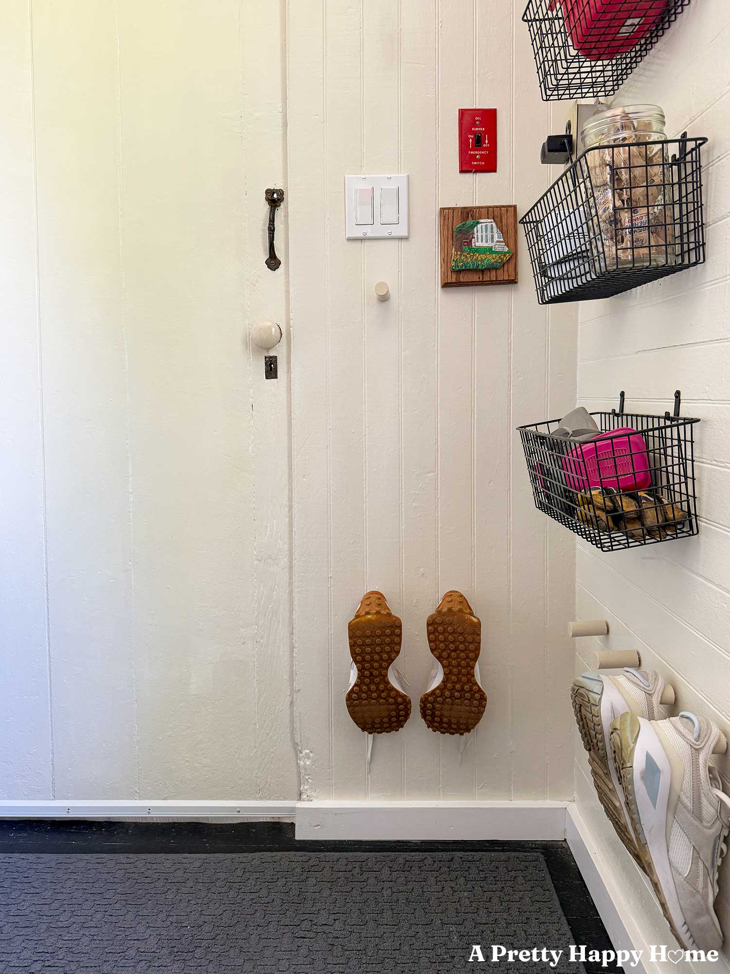 Utility room organized with metal wire baskets on the shelves to hold protective gear and laundry detergent. Shoe pegs on the wall hold tennis shoes. A small painting of a house decorates the wall next to the light switch.
