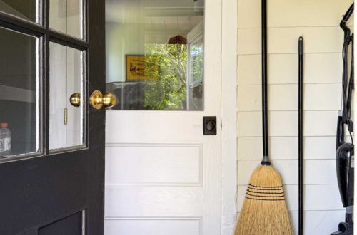black half lite door opening into a utility room space where a broom, mop, and vacuum hang on a wall next to a white door. There's a small painting hanging above the mop.
