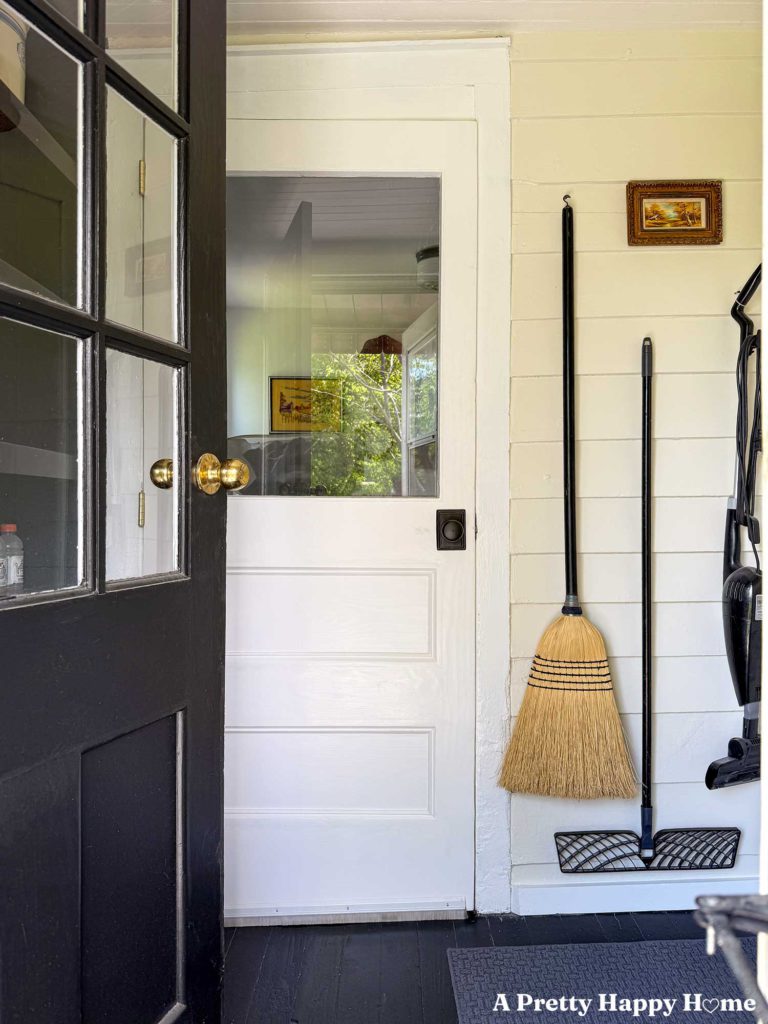 black half lite door opening into a utility room space where a broom, mop, and vacuum hang on a wall next to a white door. There's a small painting hanging above the mop.