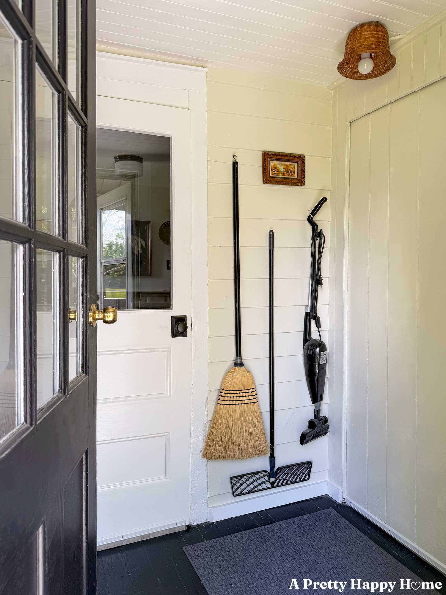 black door opening to a utility room where a broom, mop, and vacuum hang on a wall next to a white half lite door. There's a ceiling fixture with a basket for a shade.