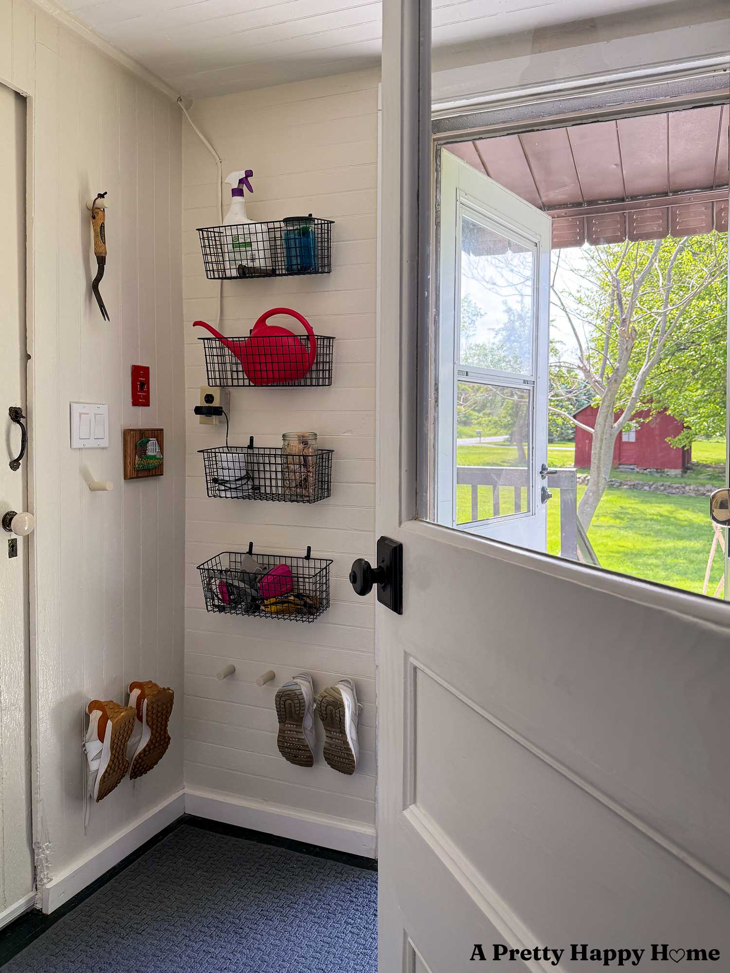 Utility room organized with metal wire baskets on the shelves to hold protective gear and laundry detergent. Shoe pegs on the wall hold tennis shoes. A small painting of a house decorates the wall next to the light switch. A door is open to the outside and there's a red barn in the distance.