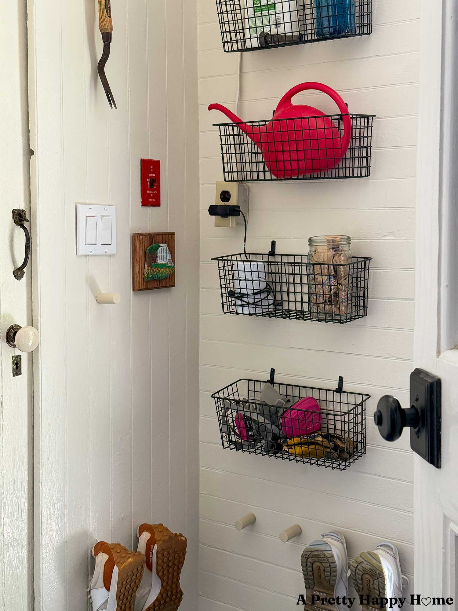 Utility room organized with metal wire baskets on the shelves to hold protective gear and laundry detergent. Shoe pegs on the wall hold tennis shoes. A small painting of a house decorates the wall next to the light switch.