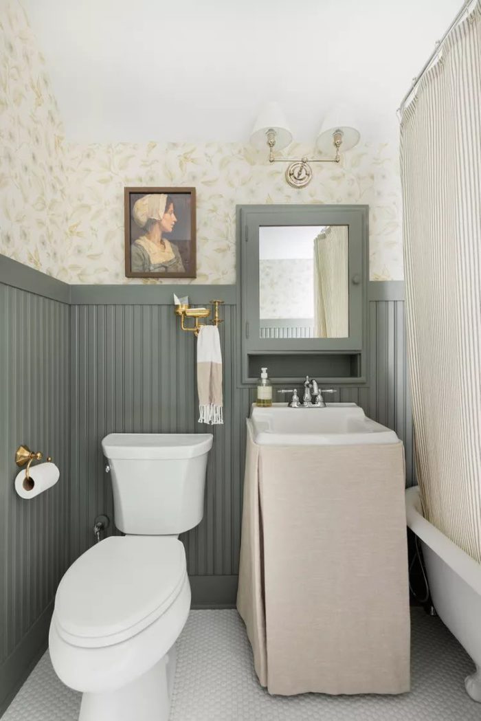 small bathroom with green wainscoting. Built into the wainscoting is medicine cabinet mirror of the same tree color with a little shelf underneath. The small sink has a linen skirt covering the bottom of it. Design by Bria Hammel Interiors
