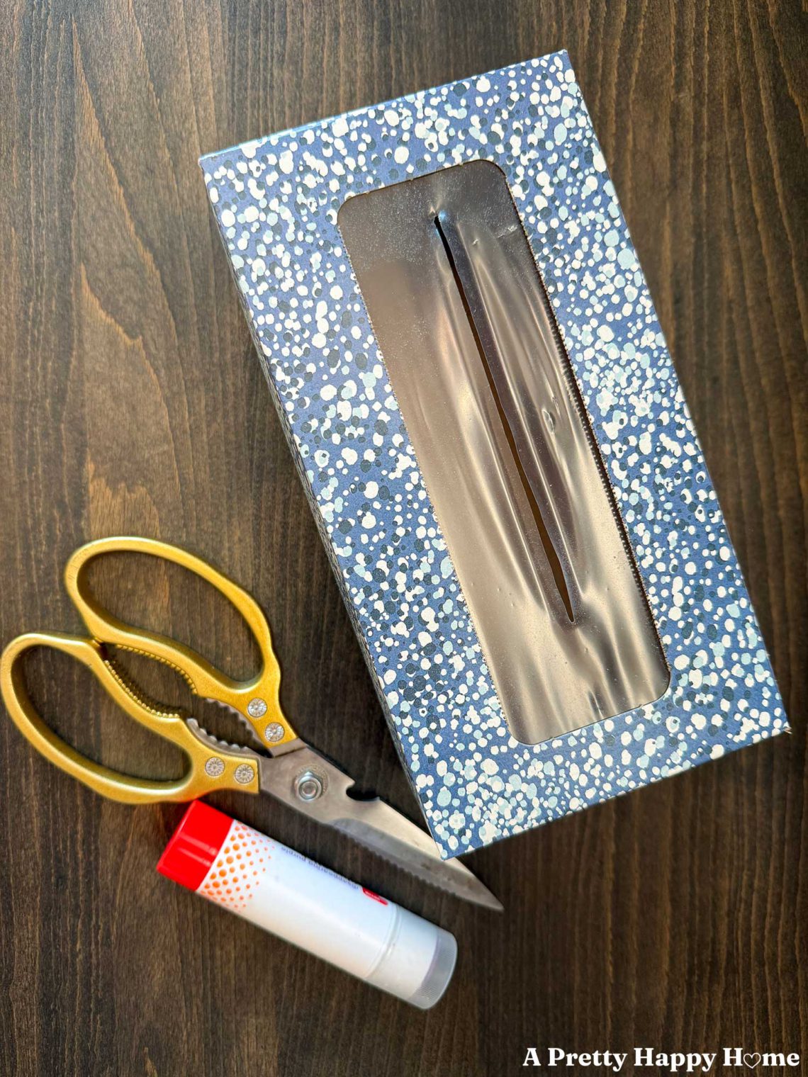 empty tissue box with a pretty blue print sitting next to gold-handled scissors and a glue stick in preparation to epicycle the tissue box cardboard into cute crafts