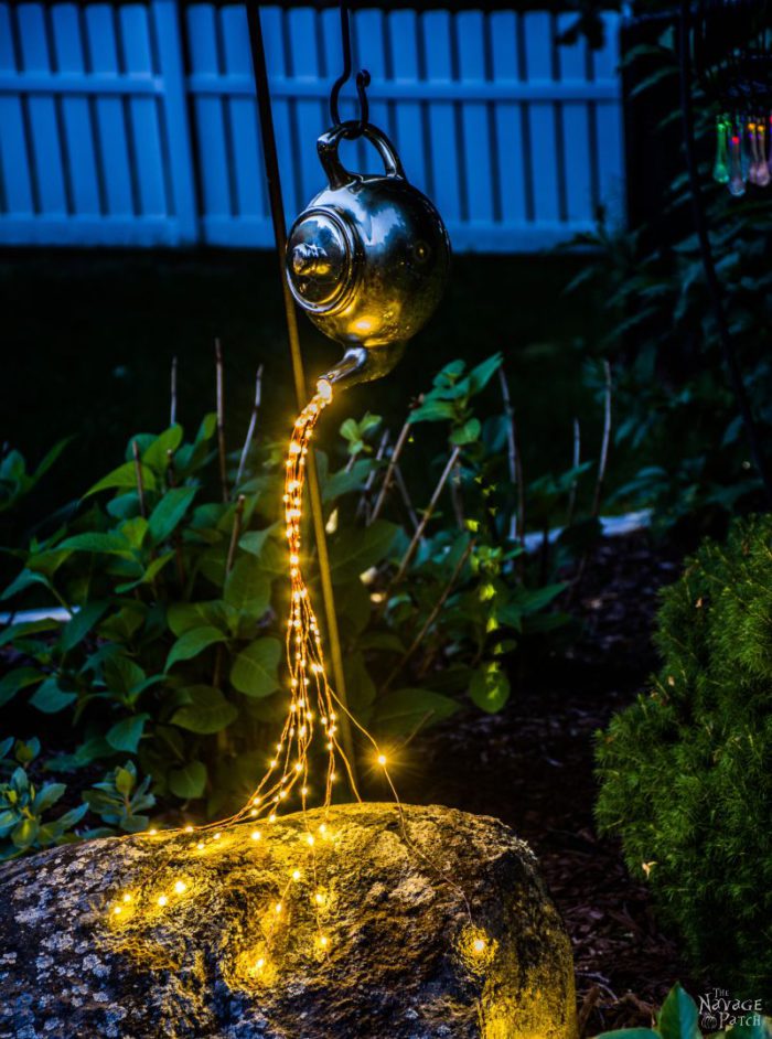 A teapot in a garden at night spilling light out of the spot with multiple strands of solar lights