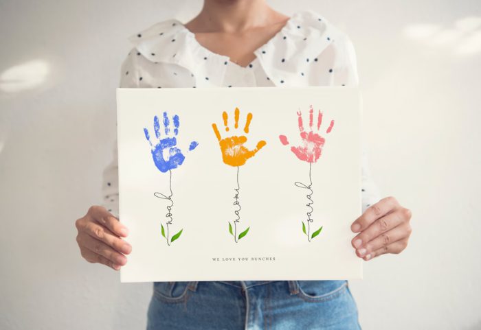 woman wearing black and white polka dotted blouse and jeans holding flower art made with toddler handprints for the flower part and their name written in script for the stem as a mother or father's day gift