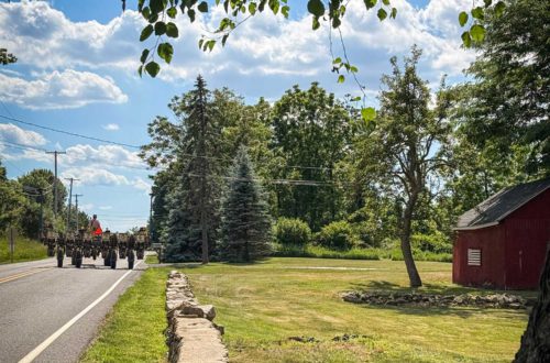 tractor driving down a paved road. There's a low rock wall on the property to the right of the road, green grass, and a red barn with an American flag hanging on the side.