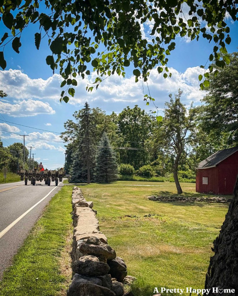 tractor driving down a paved road. There's a low rock wall on the property to the right of the road, green grass, and a red barn with an American flag hanging on the side.