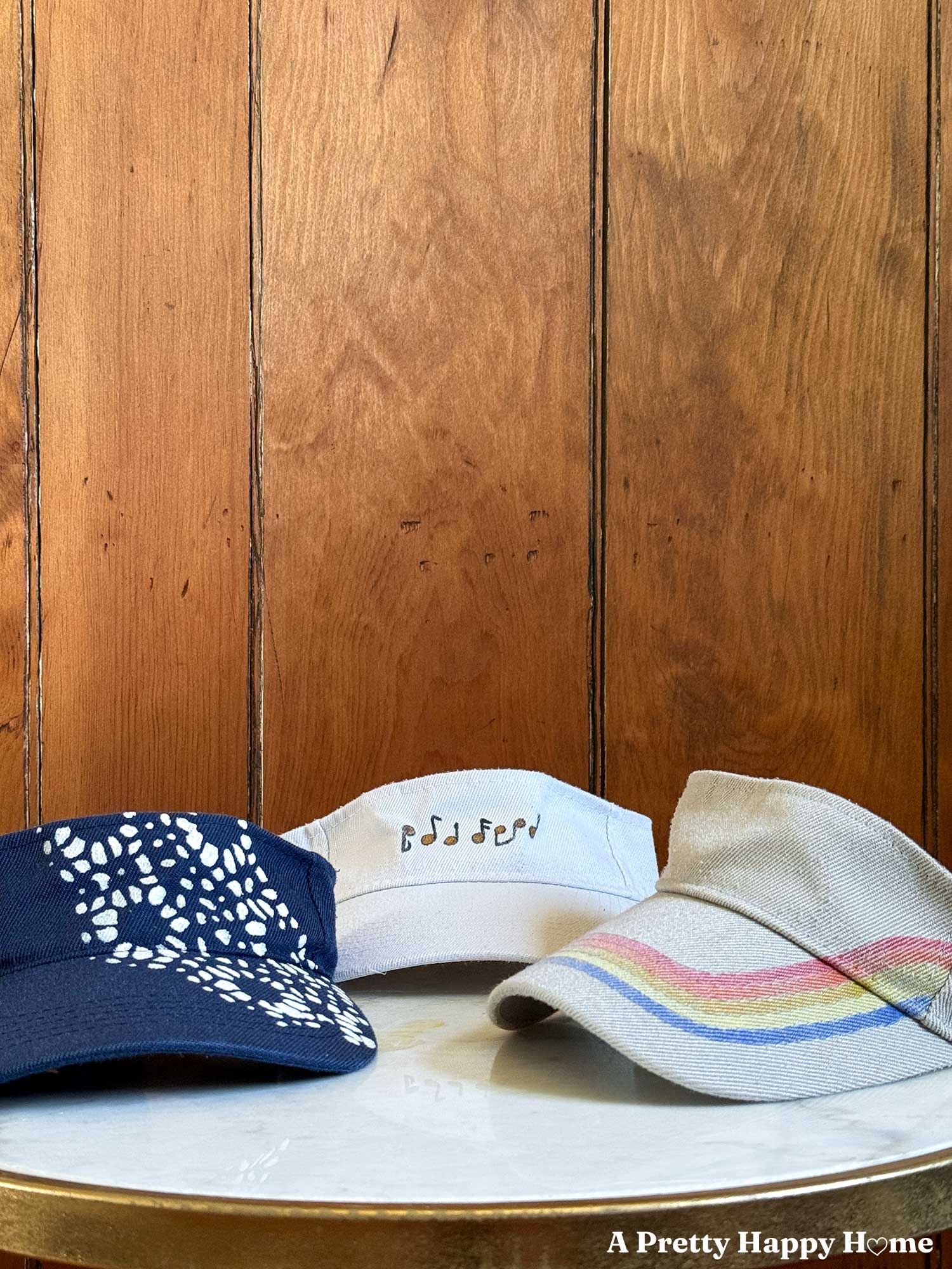 three visors sitting on a table. a blue one with a white pattern painted on an angle, a great one with retro stripes, and a white one with gold music notes painted on the headband