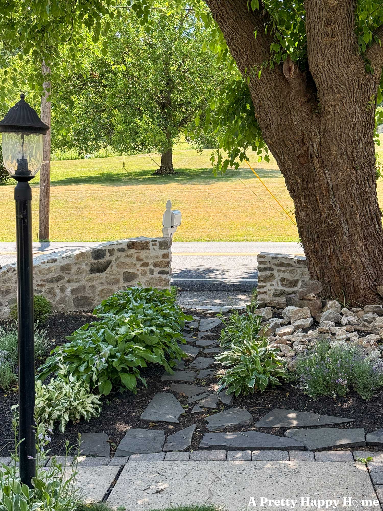 two curved and sloped fieldstone walls that flank a flagstone path. There's a elm tree in the background and black lamppost in the foreground.