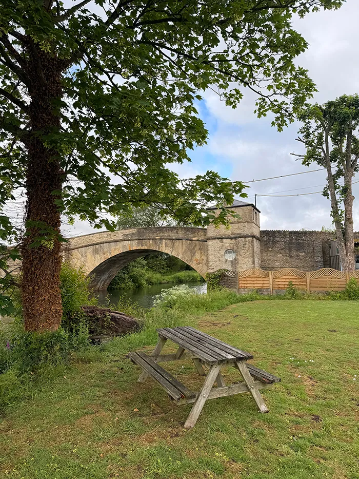 bridge in the cotswolds near a fence with curved-topped lattice panels. there is green space and and old picnic table sitting under a tree in the foreground