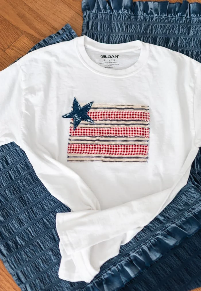 white t-shirt with patchwork flag made with strips of alternating red and white fabric with a blue fabric star over the top left corner
