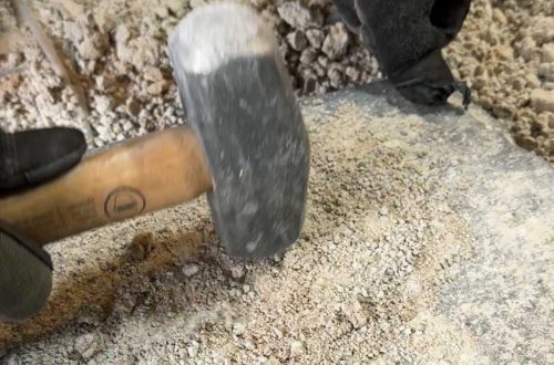 man holding a mini sledge hammer and pounding bits of old lime mortar back into sand against a piece of flagstone