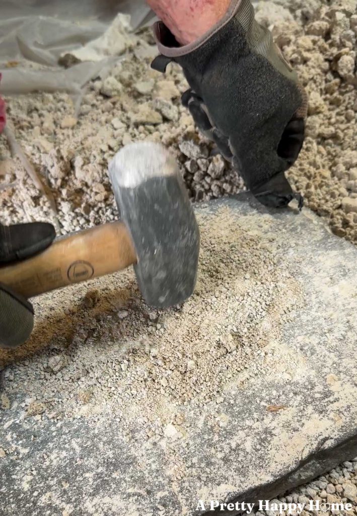man holding a mini sledge hammer and pounding bits of old lime mortar back into sand against a piece of flagstone