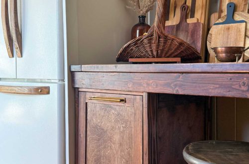 white fridge with wood handles next to a rustic table with a pull out trash cabinet built underneath it and a collection of cutting boards on top of the table