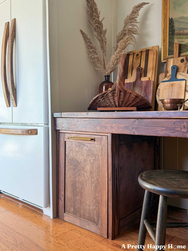 white fridge with wood handles next to a rustic table with a pull out trash cabinet built underneath it and a collection of cutting boards on top of the table