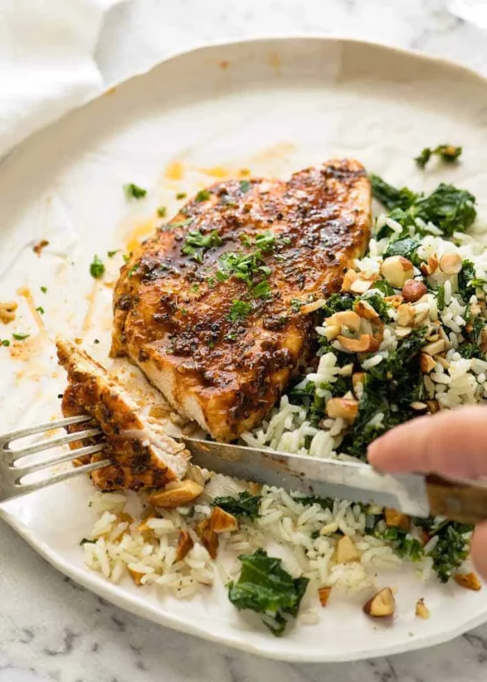 person using a knife and fork to cut oven baked chicken breast next to a mound of rice and greens