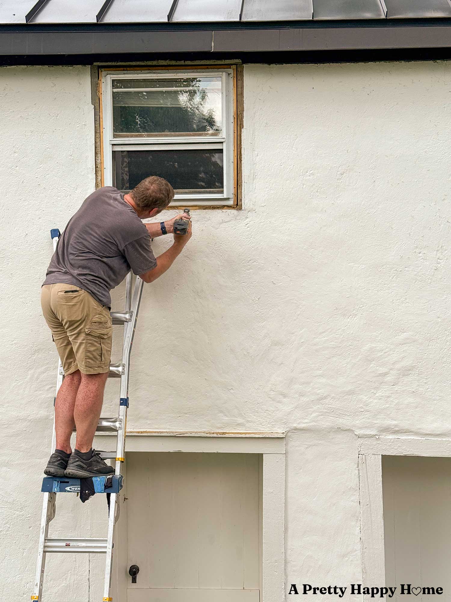 man on a ladder repairing the window trim on a white stone building with a black metal roof