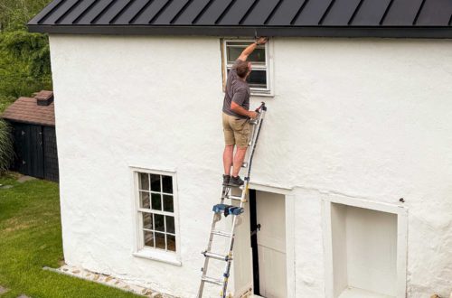 man standing on a ladder to repair window trim on a carriage house building with white masonry render and a black metal roof