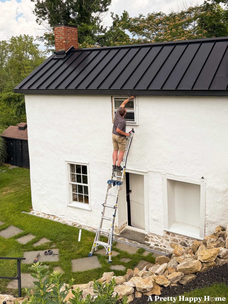 man standing on a ladder to repair window trim on a carriage house building with white masonry render and a black metal roof
