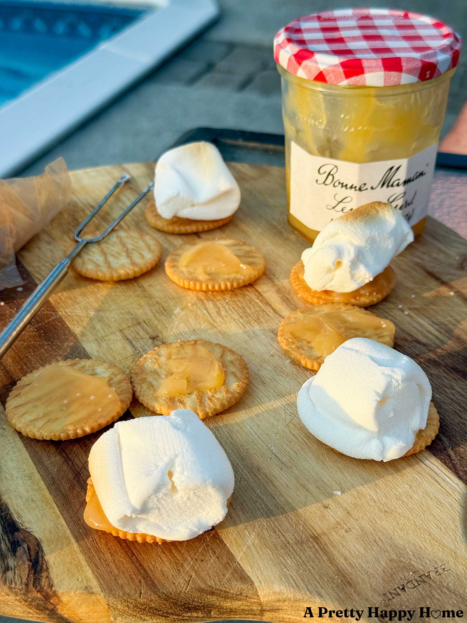 wood cutting board holding the makings of lemon curd s'mores, ritz crackers, lemon curd, melted marshmallows all next to a jar of bonne maman lemon curd.