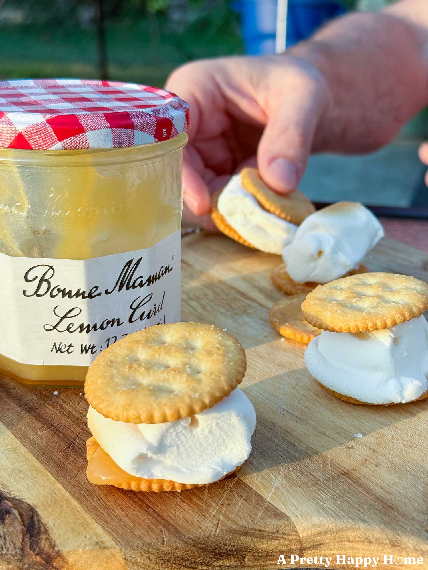 man's hand reaching for a lemon curd s'more which tastes like lemon meringue pie and the s'mores are sitting next to a bonne maman jar of lemon curd.
