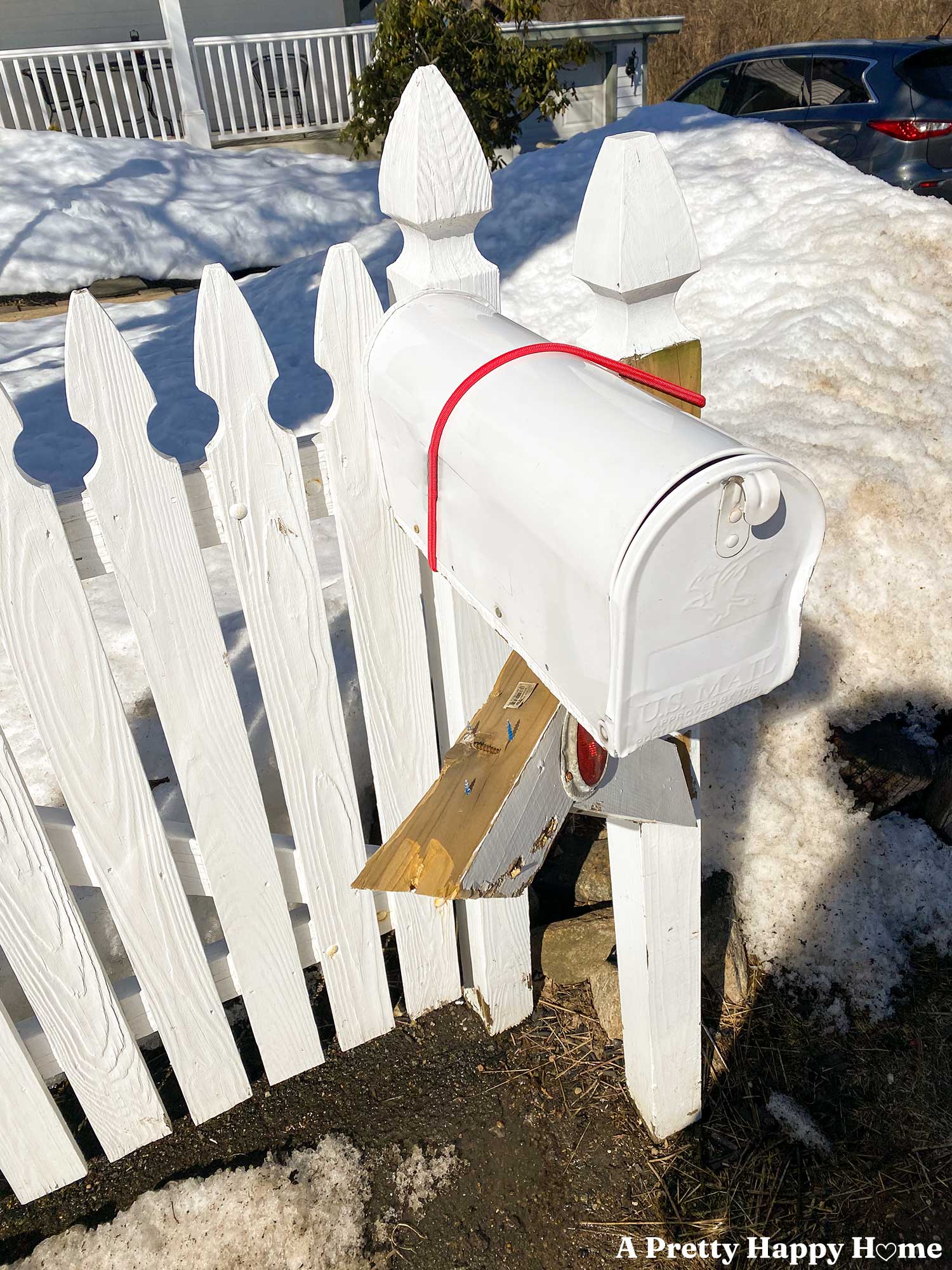 sad looking white mailbox on a wood posts that has been hit by the snowplow and held together with a red bungee cord. Snow is piled up in the background.