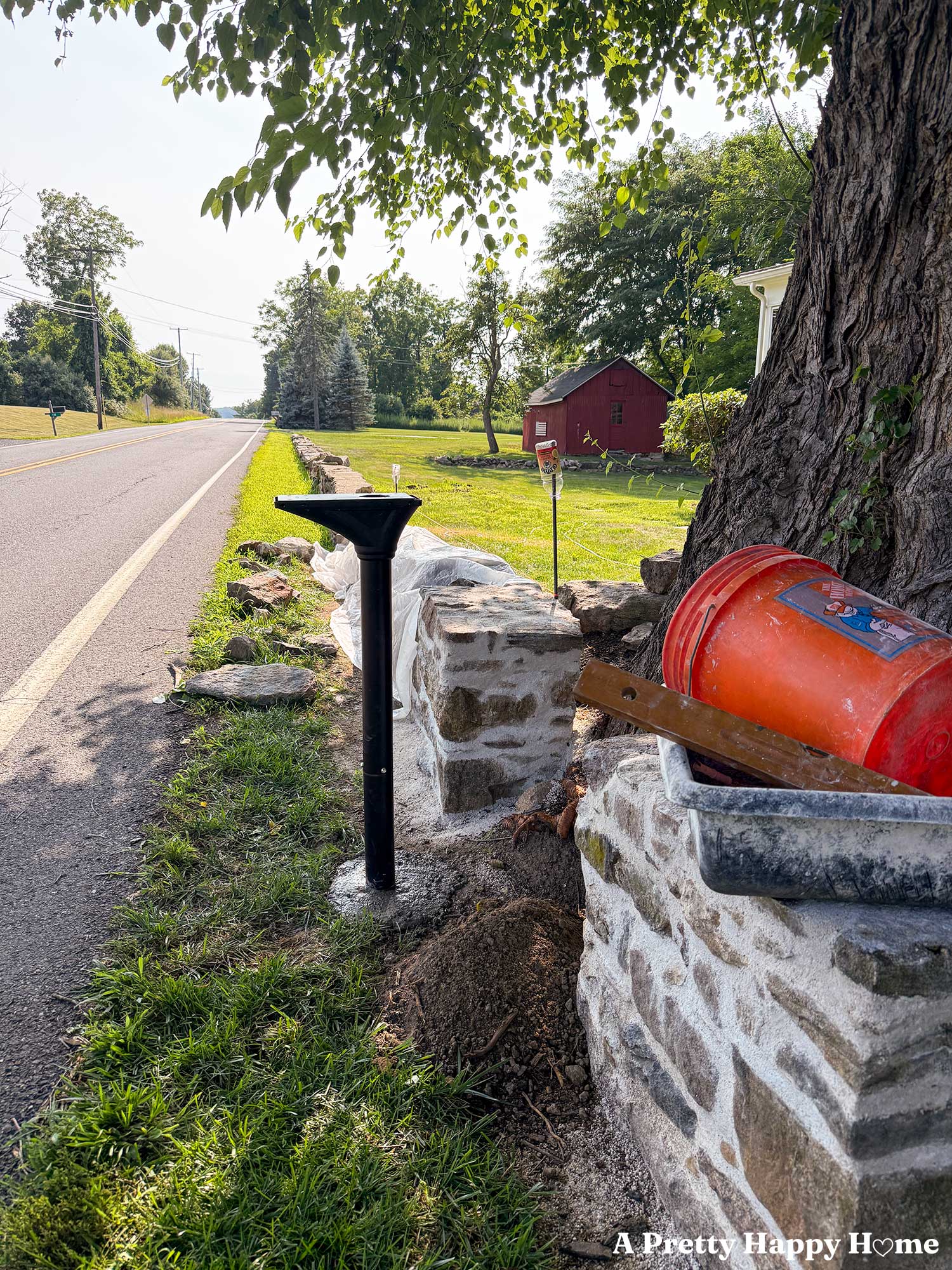 metal mailbox post in rubbed bronze drying in concrete while it waits for the mailbox itself to be installed. Low stone wall in the background is under construction.