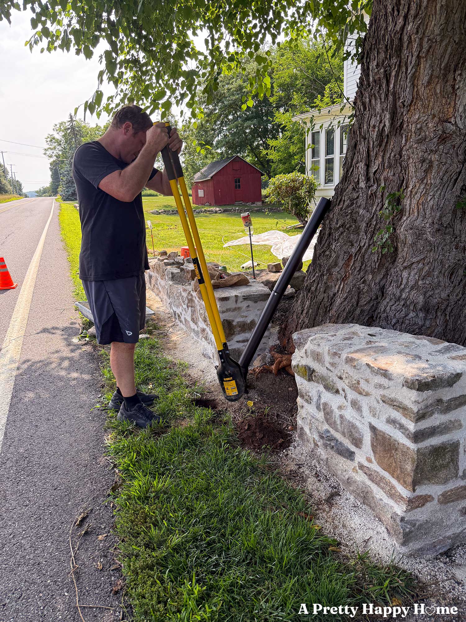 man wearing black shirt and shorts digging a hole for a mailbox post with a yellow-handled post hole digger. On either side of the hole is a low stone wall under construction.