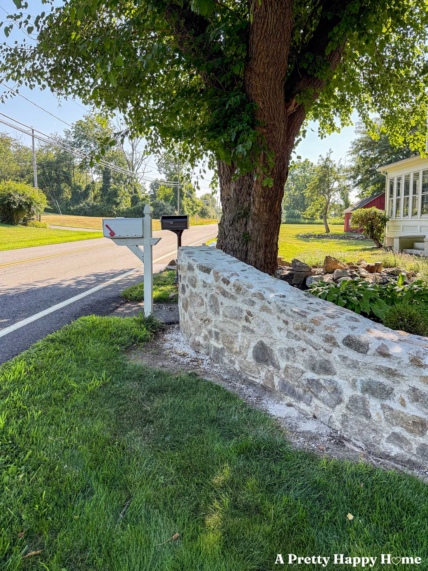 side by side comparison of an old white mailbox next to a new oil rubbed bronze mailbox that are both installed in front of a fieldstone wall and an elm tree