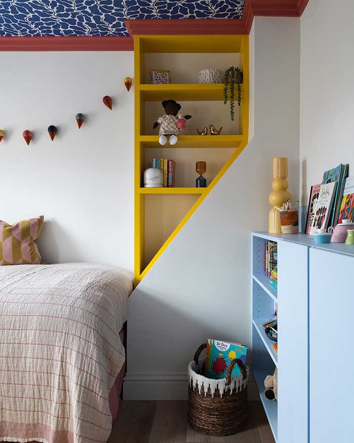 Children's room with an asymmetrical bookcase built into a quirk in the room's framing. The bookcase is painted yellow with red ceiling trim.