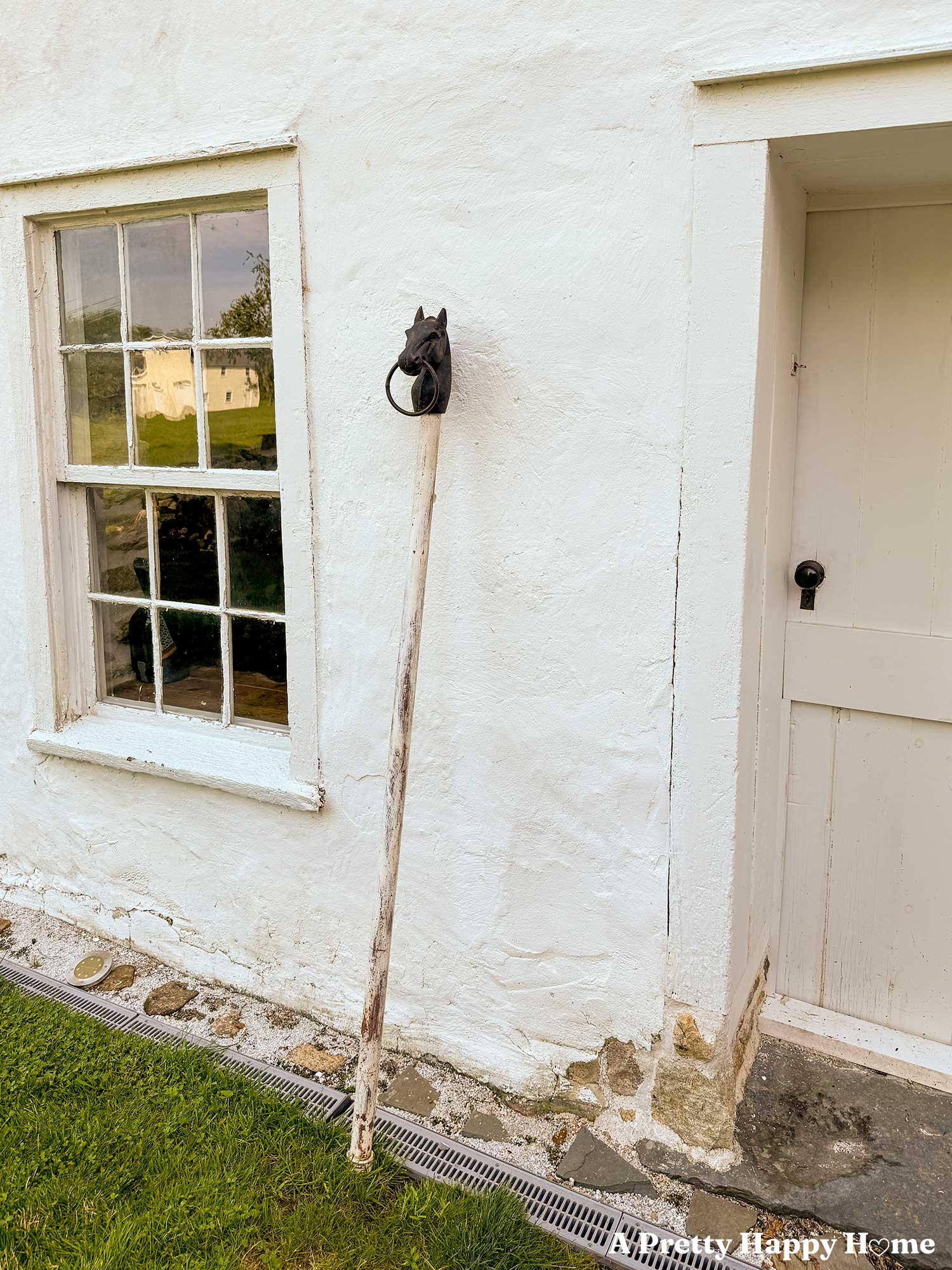 antique cast iron horse head hitching post sitting on top of a white post that will be buried in the ground and painted black to match the finial. The hitching post is leaning against a white carriage house with a six over six antique window.