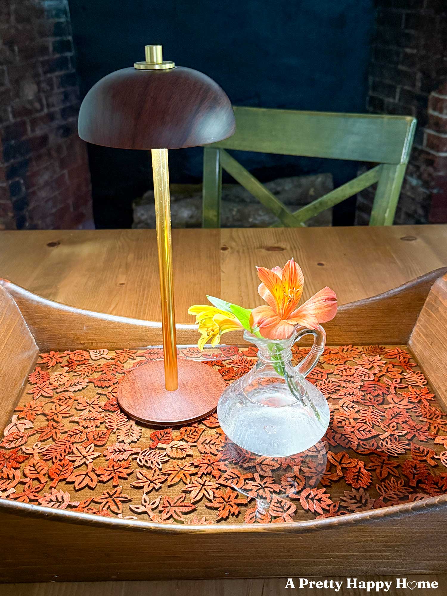 Fall-Themed Resin Tray With Wood Leaf Cutouts the tray is wood and lighter colored wood leaf cutouts line the bottom of the tray and look like they are floating in the resin. A tiny glass vase with orange flowers sits on the tray with a brown and gold lamp.