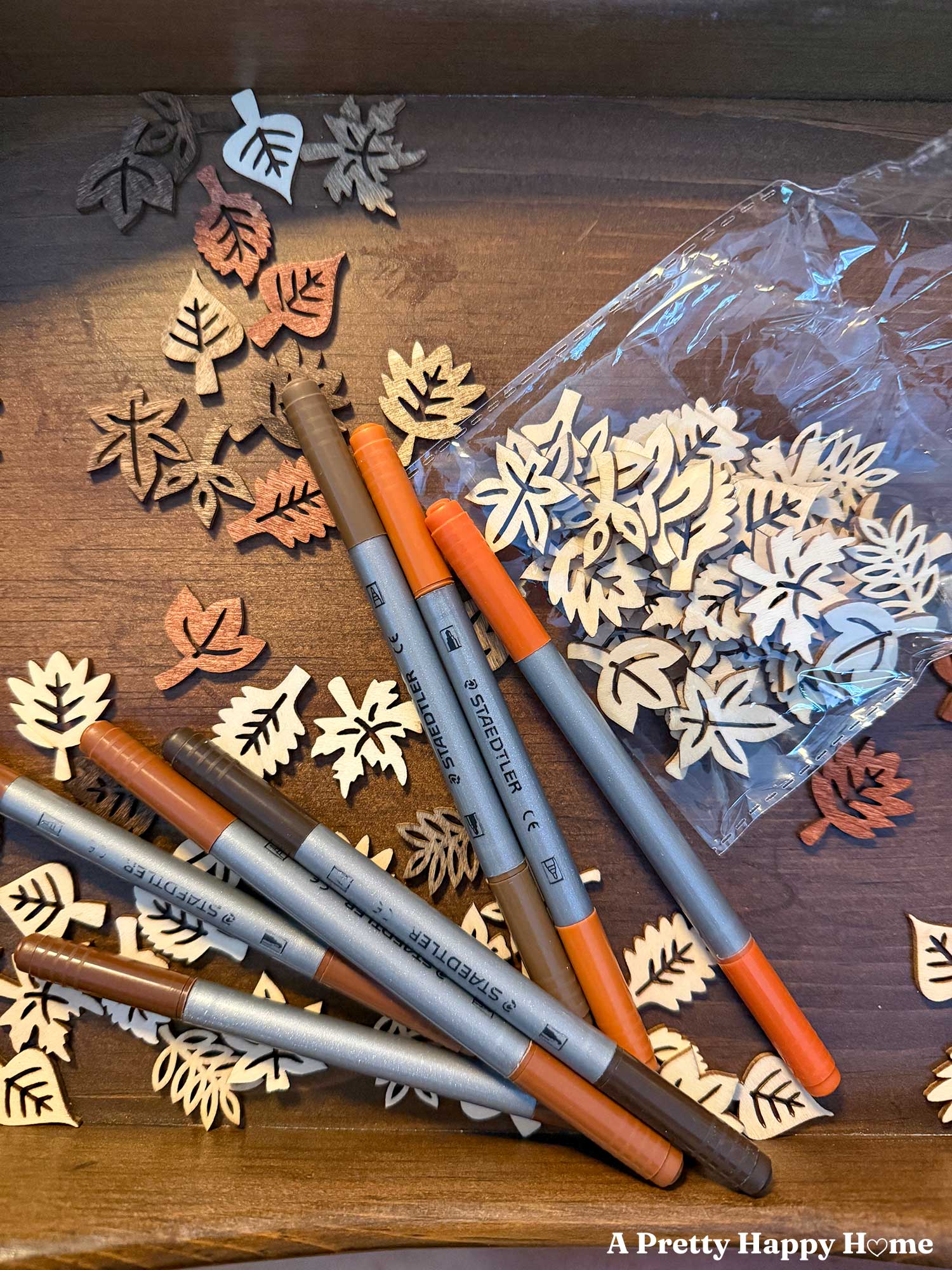 The making of a Fall-Themed Resin Tray With Wood Leaf Cutouts. the tray is wood and lighter colored wood leaf cutouts that will line the bottom of the tray and be covered in resin are being colored with permanent markers in brown and orange colors.