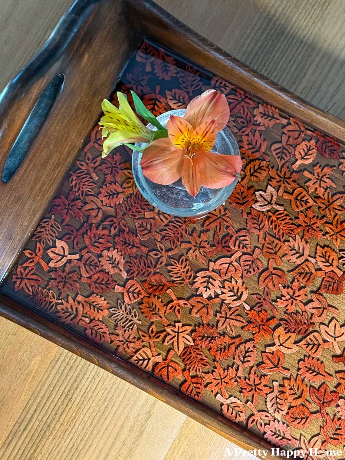 Fall-Themed Resin Tray With Wood Leaf Cutouts the tray is wood and lighter colored wood leaf cutouts line the bottom of the tray and look like they are floating in the resin. A tiny glass vase with orange flowers sits on the tray.