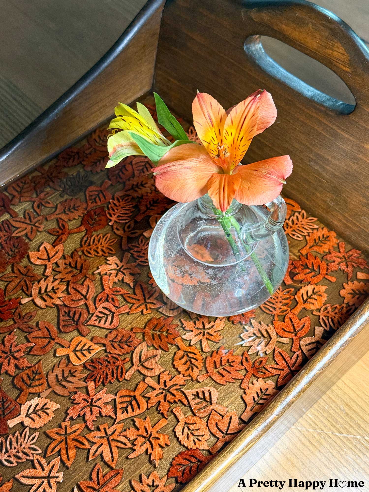 Fall-Themed Resin Tray With Wood Leaf Cutouts the tray is wood and lighter colored wood leaf cutouts line the bottom of the tray and look like they are floating in the resin. A tiny glass vase with orange flowers sits on the tray.