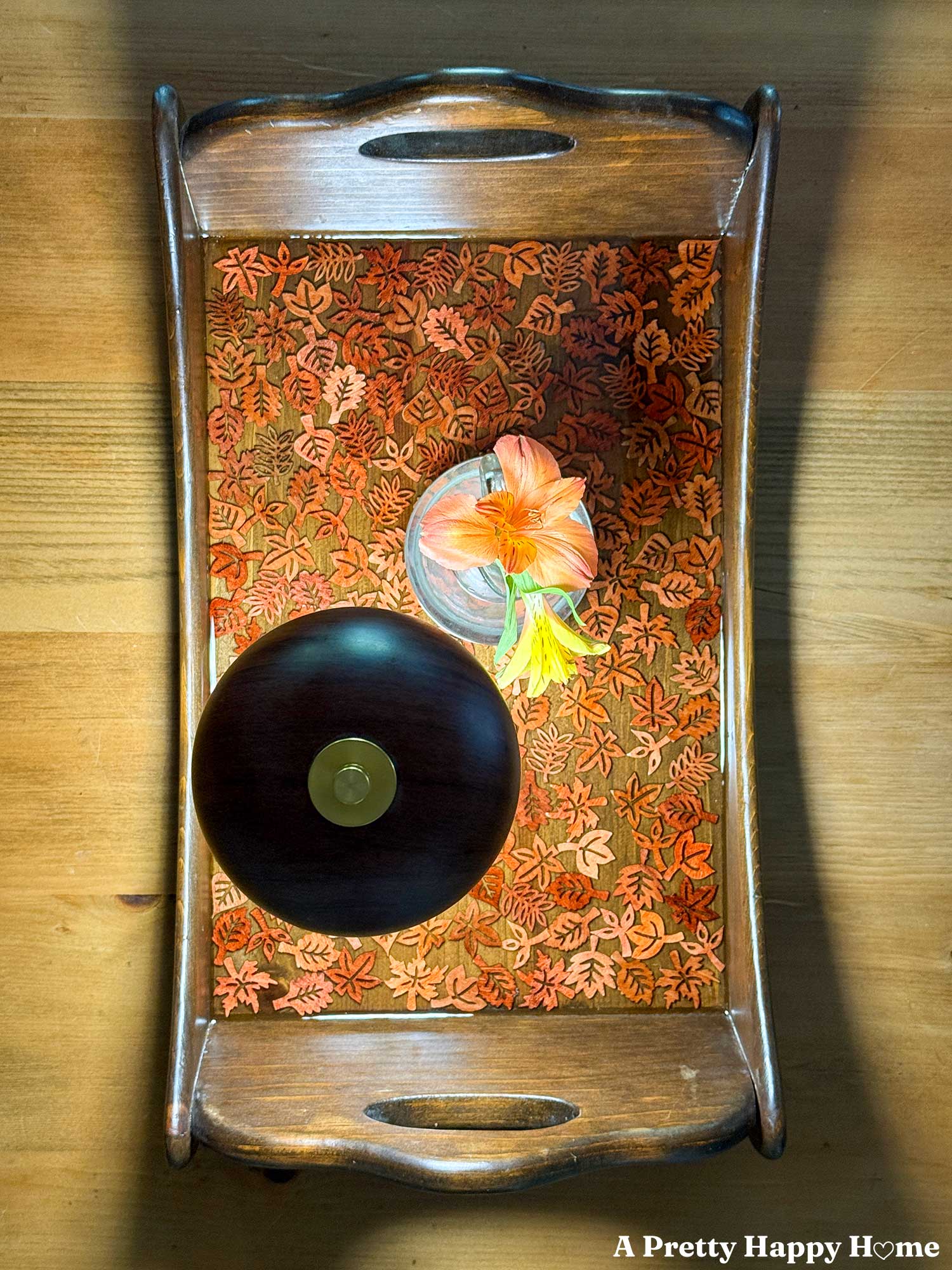 Fall-Themed Resin Tray With Wood Leaf Cutouts the tray is wood and lighter colored wood leaf cutouts line the bottom of the tray and look like they are floating in the resin. A tiny glass vase with orange flowers sits on the tray next to a brown and gold lamp.