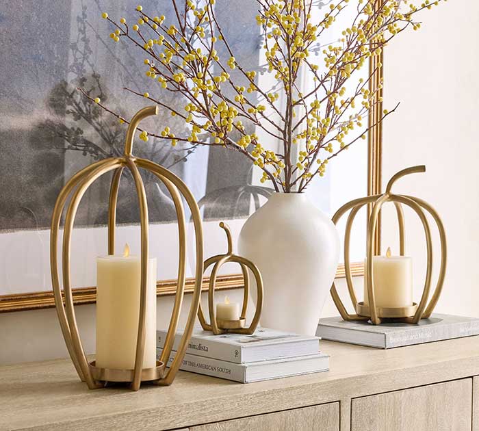 handcrafted brass pumpkin candle holders where the flameless candle sits inside the pumpkin. Trio of them are sitting on a credenza next to a white vase filled with branches.