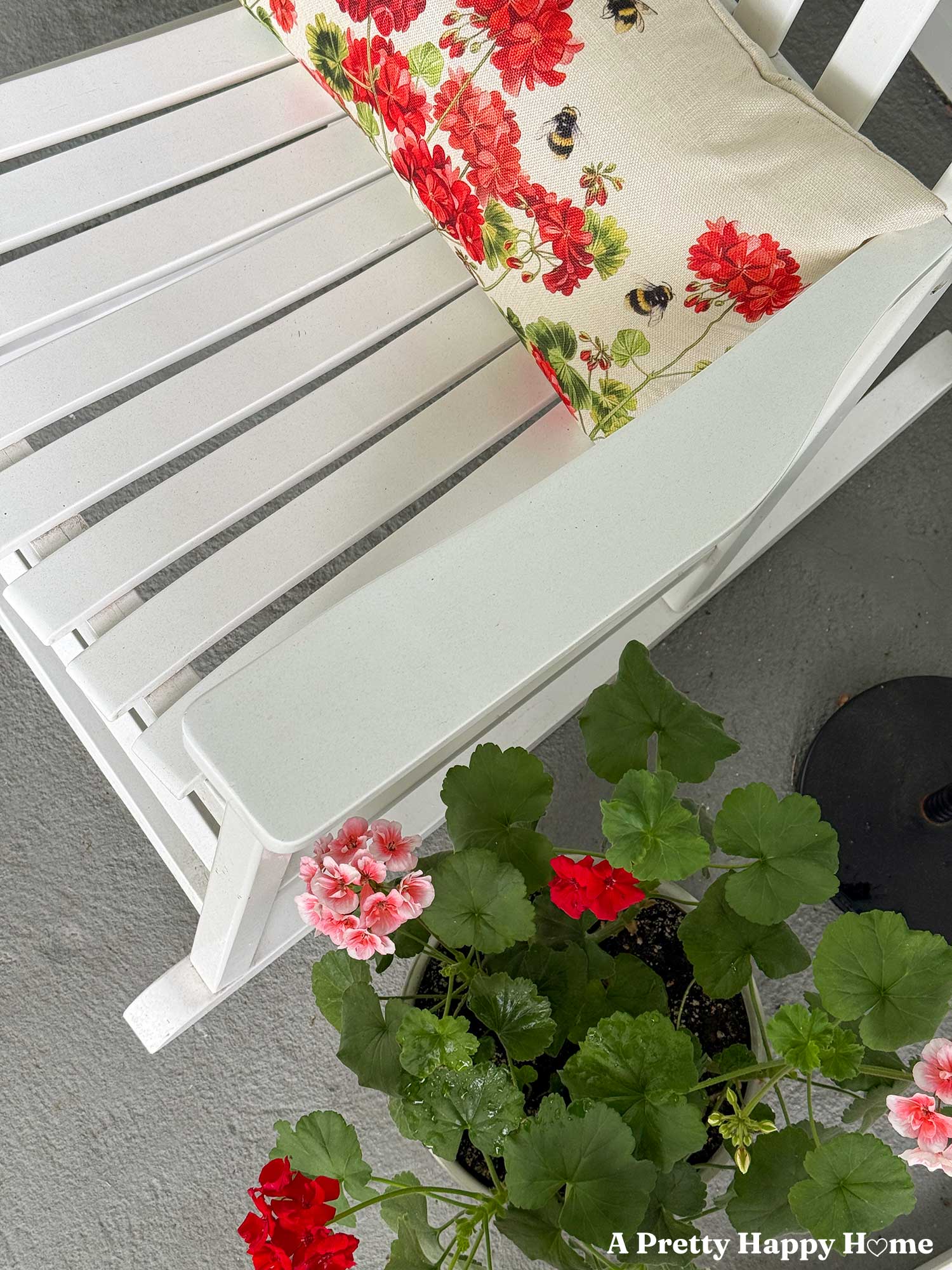 looking down at a white rocking chair with a geranium-printed pillow sitting next to a planter growing pink and red geraniums