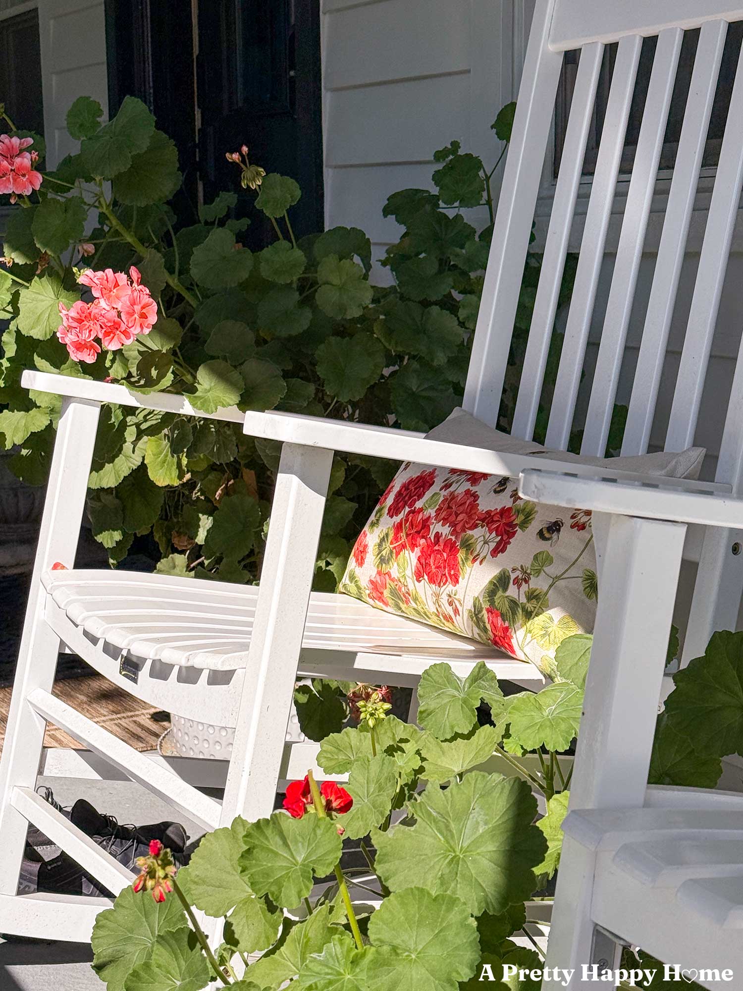 planters of pink and red geraniums surround a white rocking chair with a geranium themed throw pillow sitting on it