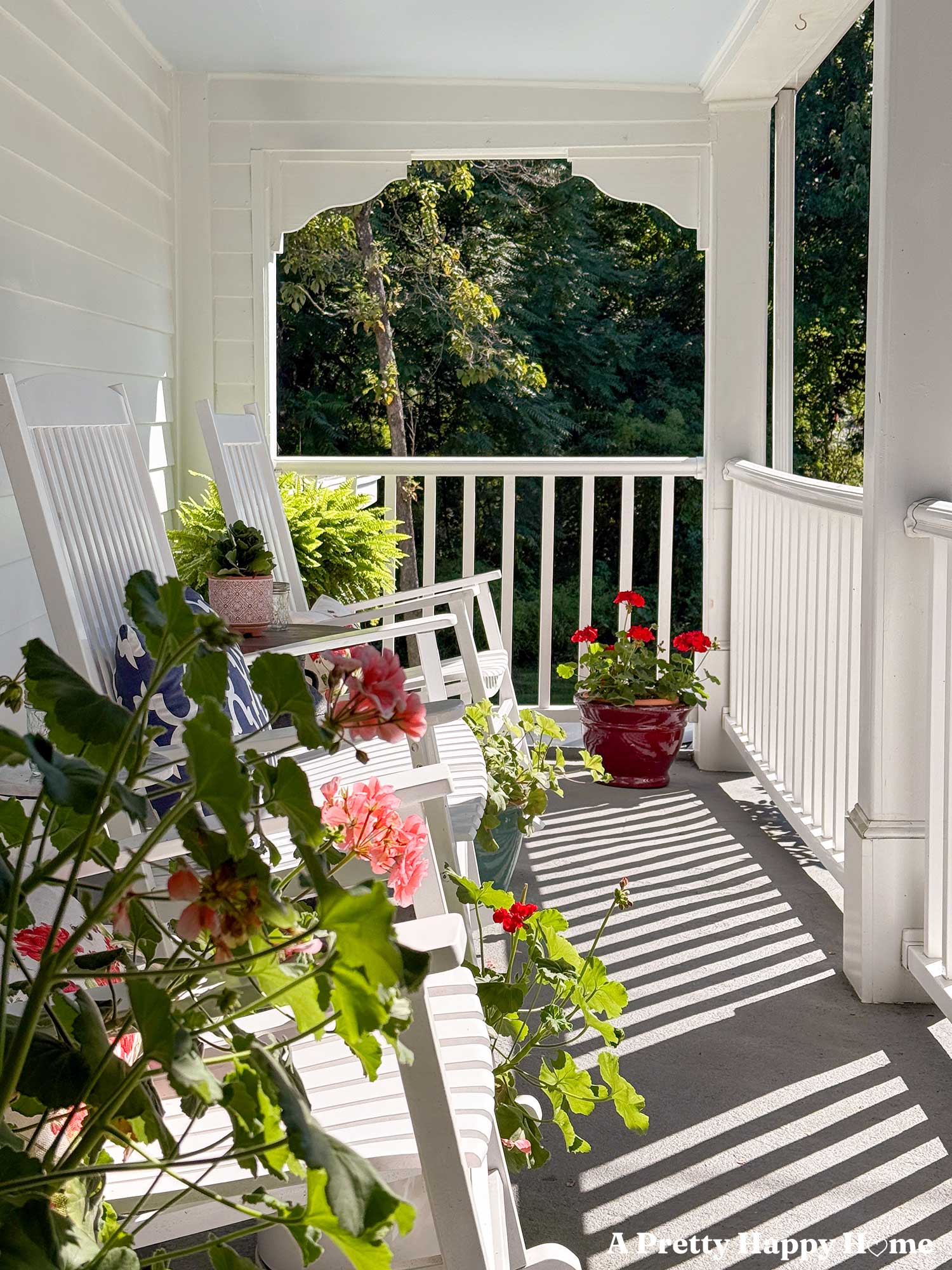 front porch with white siding, a light blue ceiling, and white railing that has white rocking chairs and large planters of blooming red and pink geraniums