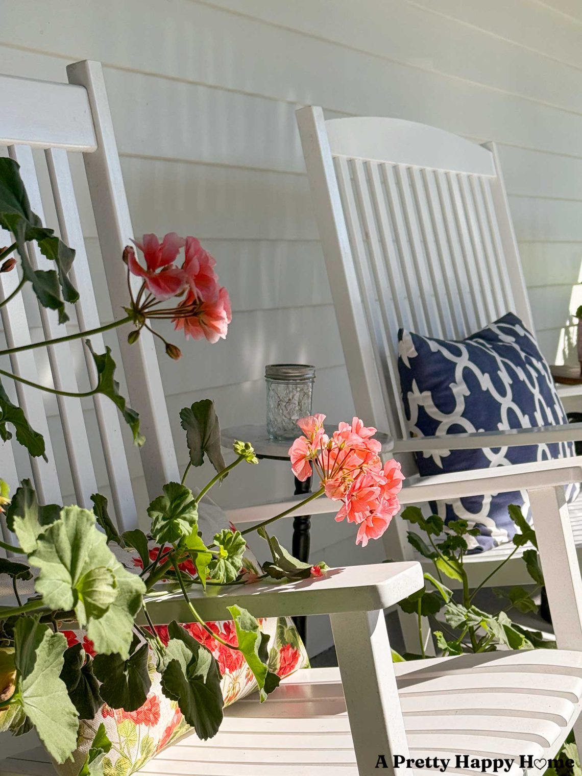Pink geraniums growing on a front porch in front of two white rocking chairs, one of which has a blue and white pillow sitting on it.