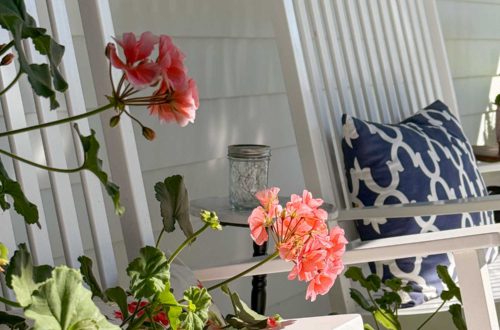 Pink geraniums growing on a front porch in front of two white rocking chairs, one of which has a blue and white pillow sitting on it.